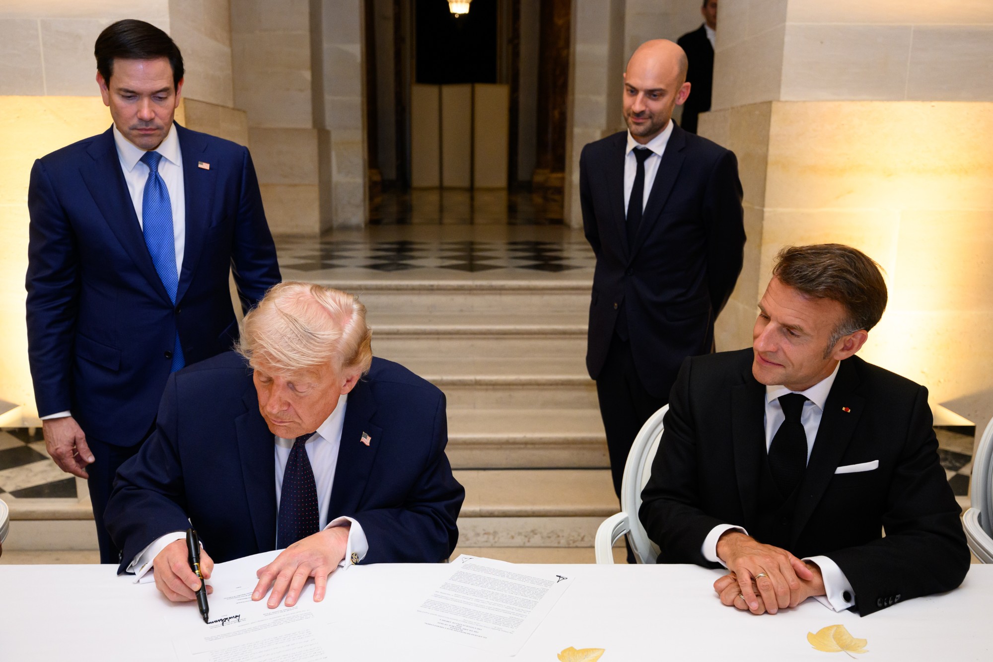 President Donald Trump signs the Islamabad Memorandum of Understanding at a table in the Palace of Versailles while Secretary of State Marco Rubio stands behind him and President Emmanuel Macron looks on
